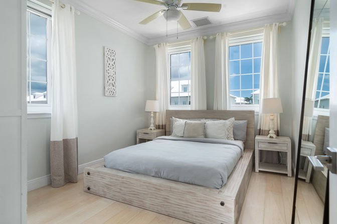 Bright coastal-style bedroom with light wood platform bed, soft gray bedding, matching nightstands and lamps, white sheer curtains, ceiling fan, and large multi-pane windows showing blue sky.