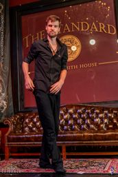 Performer in a black shirt with headset mic walking across an intimate lounge stage featuring a tufted leather sofa, patterned rug and a playing card on the floor.