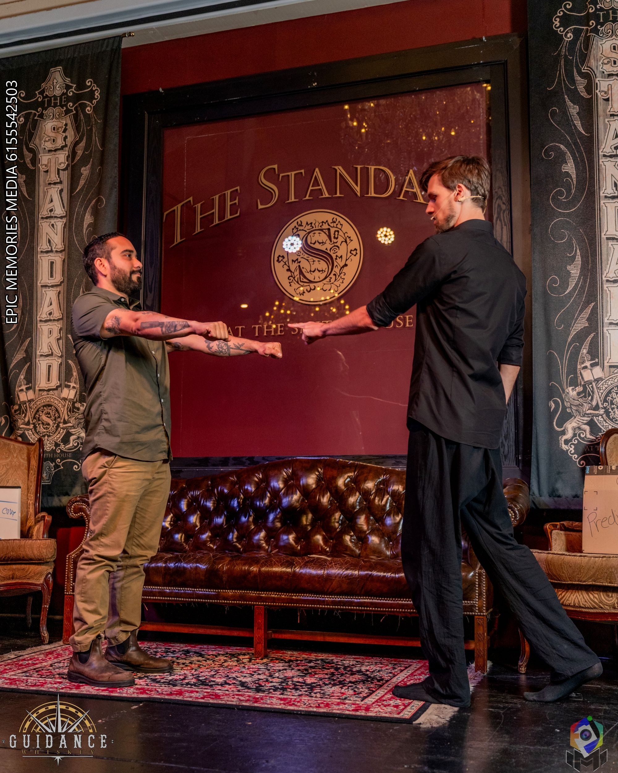 Two performers on a vintage lounge stage extend their arms in a playful mock duel or magic routine, standing before a leather sofa and ornate backdrop under warm stage lighting.
