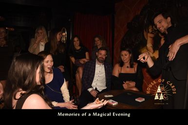 Dimly lit indoor magic show with a group of adults gathered around a table watching a magician perform a card trick, playing cards and drinks on the table, caption reads “Memories of a Magical Evening.”