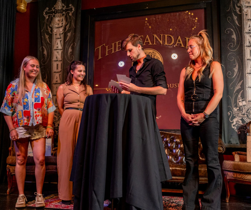Four adults on a small indoor stage at a live show: a host reading cards at a tall draped cocktail table while three participants smile, set against leather seating and an ornate lounge backdrop.