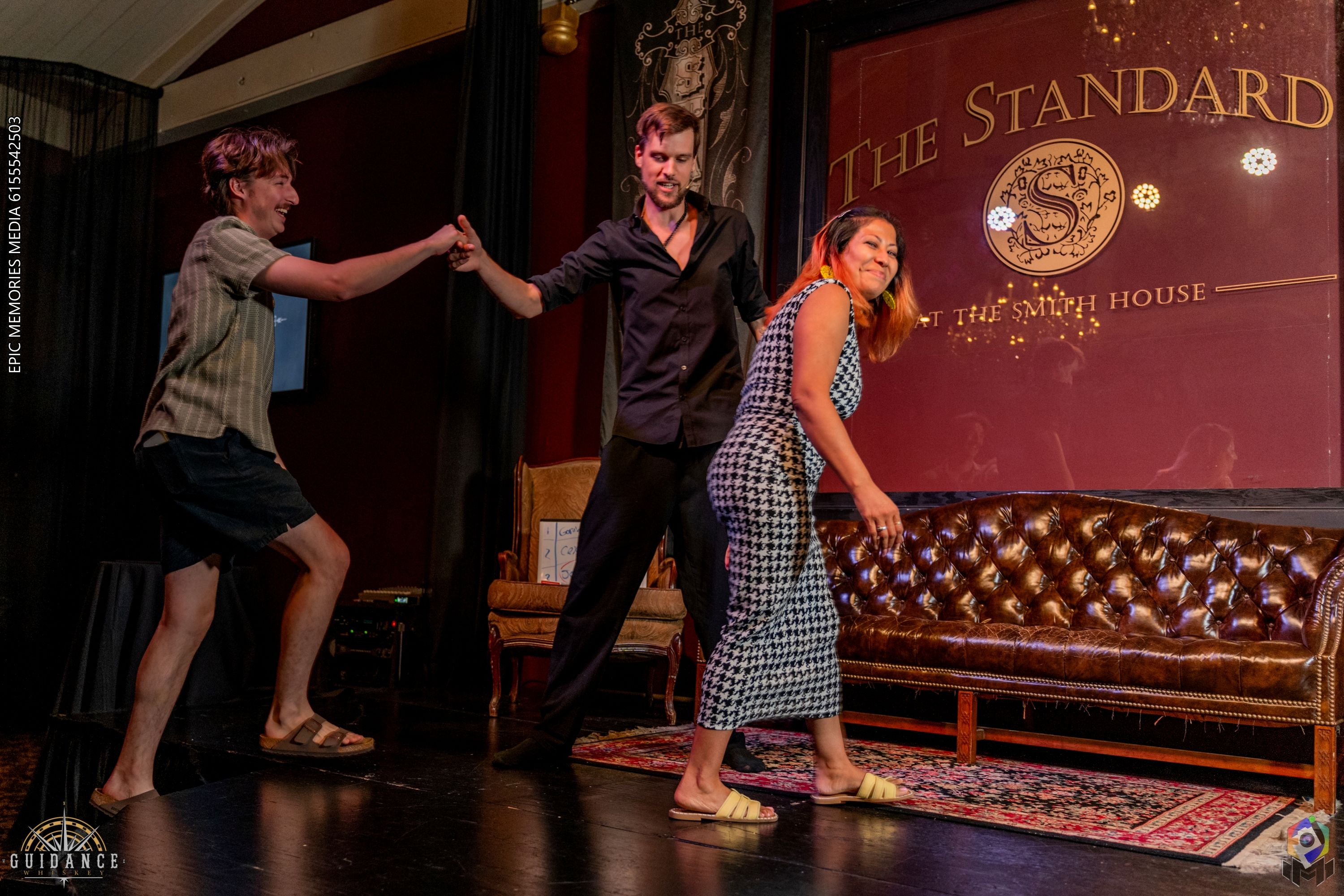 Three adults on a cozy indoor stage with a tufted leather sofa and rug — a man in shorts reaches out as a man in a black shirt steadies him, while a smiling woman in a patterned dress steps forward.