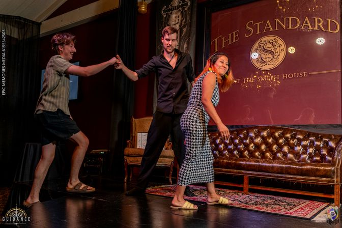 Three adults on a cozy indoor stage with a tufted leather sofa and rug — a man in shorts reaches out as a man in a black shirt steadies him, while a smiling woman in a patterned dress steps forward.