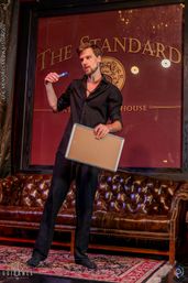 Performer in a black shirt holding a marker and small whiteboard on a cozy lounge stage with a tufted leather sofa and burgundy backdrop.
