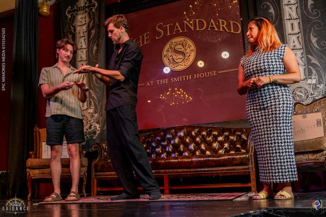 Three performers on an intimate vintage theater stage: a performer in black demonstrating a sleight-of-hand trick with a volunteer while a woman in a houndstooth dress watches beside a tufted leather sofa under warm chandelier lighting.