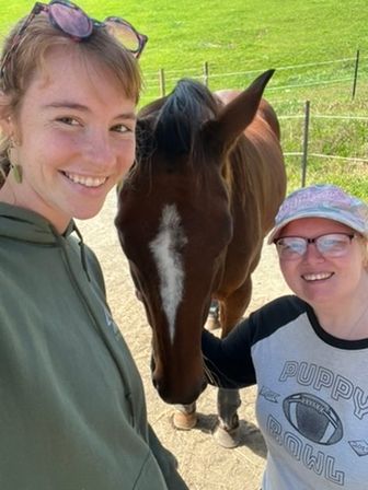 Two smiling people take a cheerful selfie with a brown horse showing a white facial blaze, standing on a sunny country path beside a green pasture and fence.