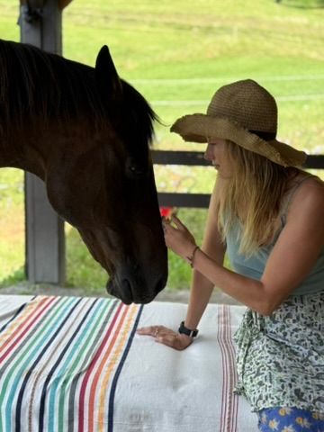 Woman in a straw sun hat offering a small red flower to a brown horse across a striped cloth-covered table on a farmhouse porch with green pasture behind