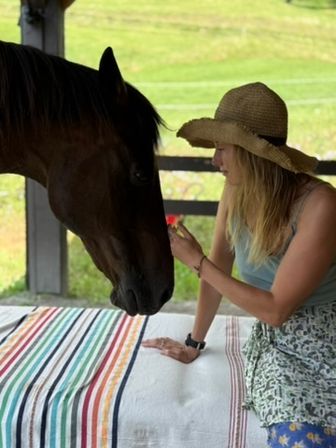 Woman in a straw sun hat offering a small red flower to a brown horse across a striped cloth-covered table on a farmhouse porch with green pasture behind