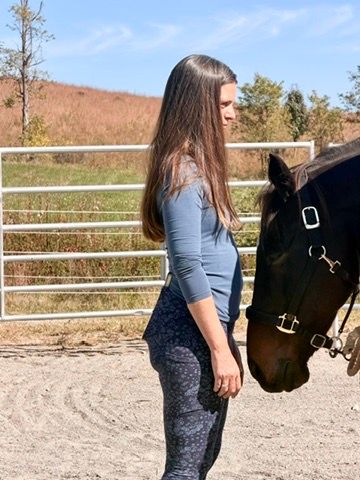 Woman with long brown hair standing beside a dark brown horse in a sunny outdoor riding arena, sandy footing, metal fence and grassy hillside in the background.