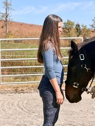 Woman with long brown hair standing beside a dark brown horse in a sunny outdoor riding arena, sandy footing, metal fence and grassy hillside in the background.