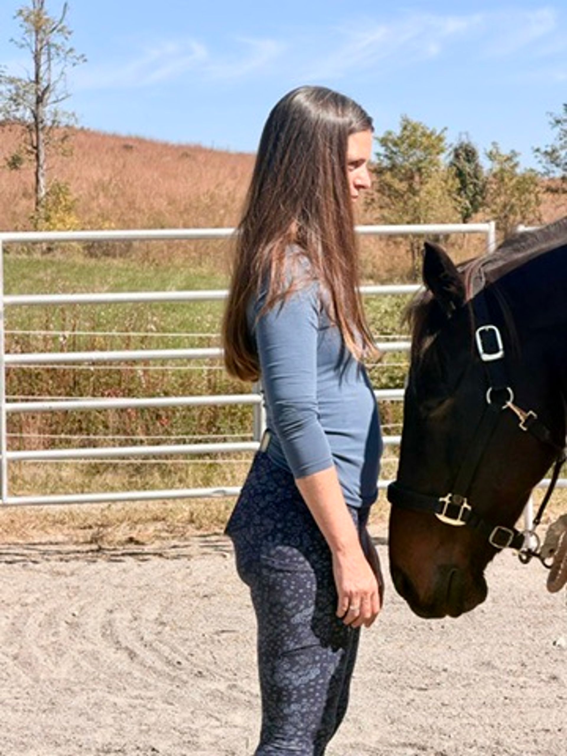 Woman with long brown hair standing beside a dark brown horse in a sunny outdoor riding arena, sandy footing, metal fence and grassy hillside in the background.