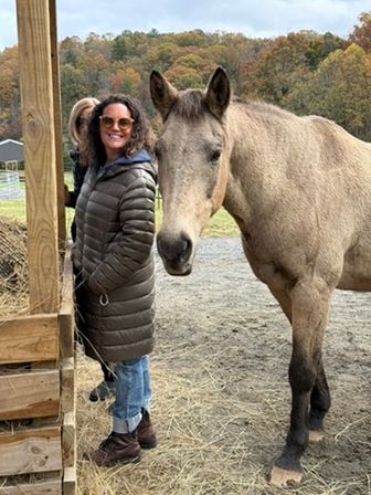 Woman in a puffer coat and sunglasses smiling beside a light-brown horse at a rural farm in autumn with colorful hillside trees in the background.