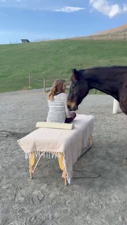 Person sitting on a covered massage table in an outdoor arena as a brown horse gently nuzzles them, green hillside and fence under a blue sky.