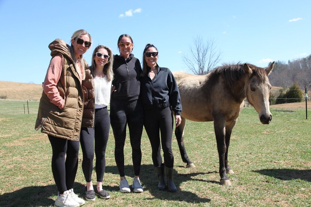 Four women in sunglasses and casual activewear smiling together in a sunny rural pasture beside a brown horse and rolling hills