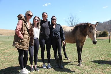 Four women in sunglasses and casual activewear smiling together in a sunny rural pasture beside a brown horse and rolling hills