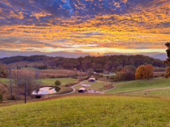 Rolling green hills and a pastoral farm with a small pond and winding country lane under a dramatic golden-orange sunset sky, autumn trees dotting the peaceful countryside