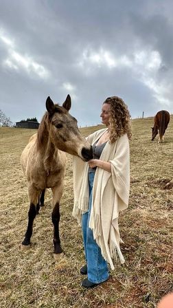 Curly-haired woman in a cream fringed shawl and jeans pets a curious light-brown horse in a grassy countryside pasture under an overcast sky, another horse grazing in the background.