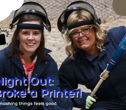 Two women in protective face shields and coveralls smile in an indoor smash room; one holds a baseball bat beside a hammer-smash icon and the overlay text "Girls Night Out: We Broke a Printer!"