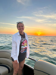Smiling person standing on a boat at sunset with a colorful sky and sun low over open water, wearing a graphic tee, white overshirt, and sunglasses perched on their head.