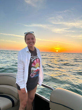 Smiling person standing on a boat at sunset with a colorful sky and sun low over open water, wearing a graphic tee, white overshirt, and sunglasses perched on their head.