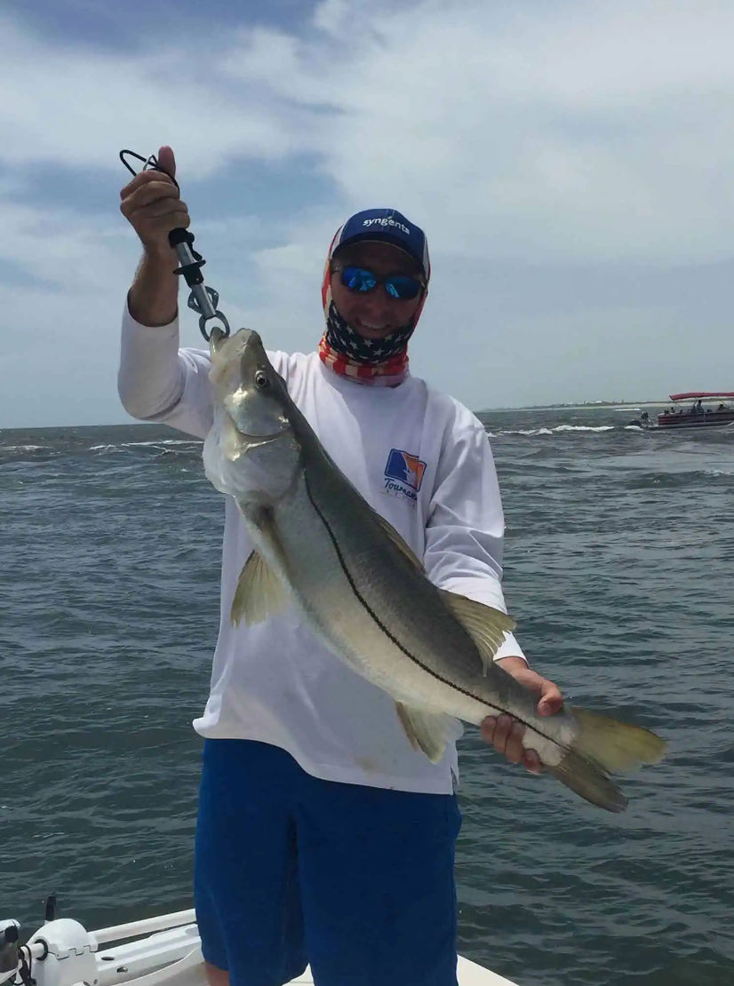 Proud angler holding a large saltwater snook with a lip gripper on a boat over choppy coastal waters under a cloudy sky