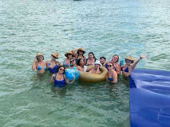 Group of friends in bikinis and sun hats smiling and holding drinks on a gold inflatable and in shallow turquoise ocean water during a sunny summer beach day.