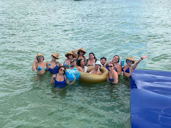 Group of friends in bikinis and sun hats smiling and holding drinks on a gold inflatable and in shallow turquoise ocean water during a sunny summer beach day.