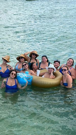 Group of friends in bikinis and sun hats smiling and holding drinks on a gold inflatable and in shallow turquoise ocean water during a sunny summer beach day.