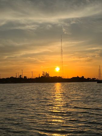 Golden sunset over a coastal marina with a tall radio tower and sailboat masts silhouetted, sun reflecting on rippled water.