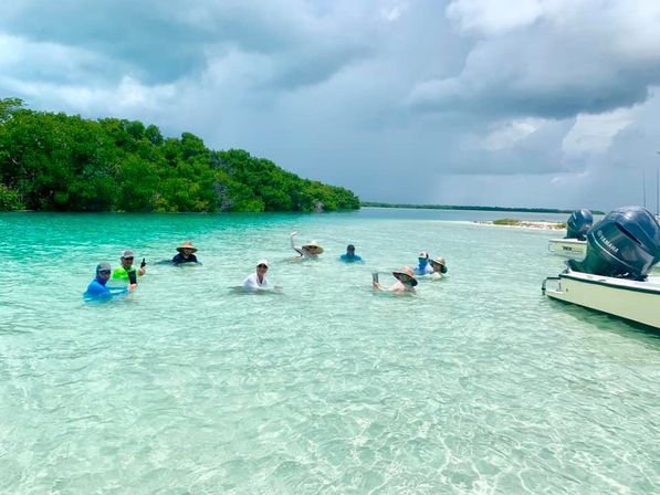 Group of people wading in shallow, crystal‑clear turquoise water near coastal mangroves with a small motorboat nearby, enjoying drinks on a tropical sandbar under a cloudy sky.