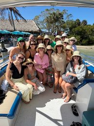 Smiling group of women in sun hats and sunglasses on a boat at a tropical dock with palm trees, a thatched-roof eatery and bright blue sky — beachside vacation vibe.