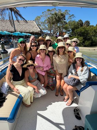 Smiling group of women in sun hats and sunglasses on a boat at a tropical dock with palm trees, a thatched-roof eatery and bright blue sky — beachside vacation vibe.