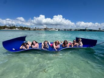 Group of friends lounging on a large blue floating mat in clear turquoise bay, holding drinks under a sunny sky with puffy clouds, palm-lined shore and a low coastal bridge in the background.
