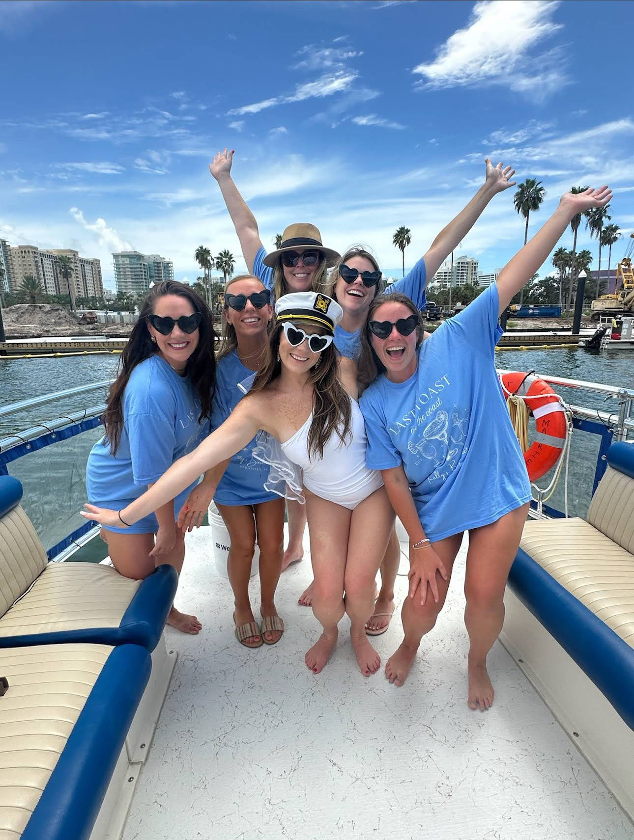 Six friends posing on a boat at a sunny palm-lined marina — five in matching blue shirts and heart-shaped sunglasses and one bride-to-be in a white swimsuit with a captain’s hat, coastal skyline and palm trees in the background.