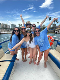 Six friends posing on a boat at a sunny palm-lined marina — five in matching blue shirts and heart-shaped sunglasses and one bride-to-be in a white swimsuit with a captain’s hat, coastal skyline and palm trees in the background.