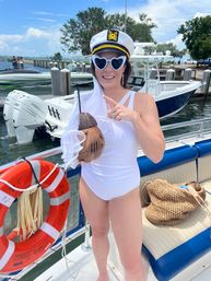 Person in a white swimsuit wearing a captain’s hat with a veil and heart-shaped sunglasses, holding a coconut drink aboard a sunlit boat at a coastal marina dock with powerboats and a life ring nearby.