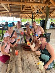Eight friends laughing and toasting colorful cocktails at a beachside tiki bar under a thatched-roof hut, seated around a weathered wooden table with palm trees and umbrellas in the sunny tropical setting.