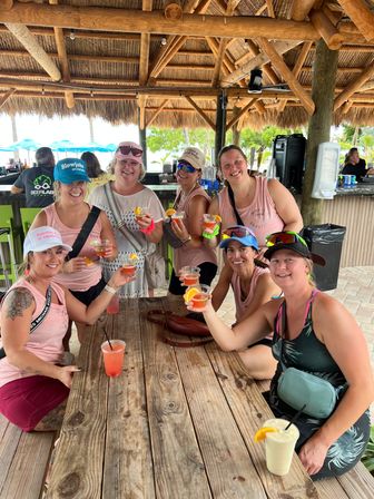 Eight friends laughing and toasting colorful cocktails at a beachside tiki bar under a thatched-roof hut, seated around a weathered wooden table with palm trees and umbrellas in the sunny tropical setting.