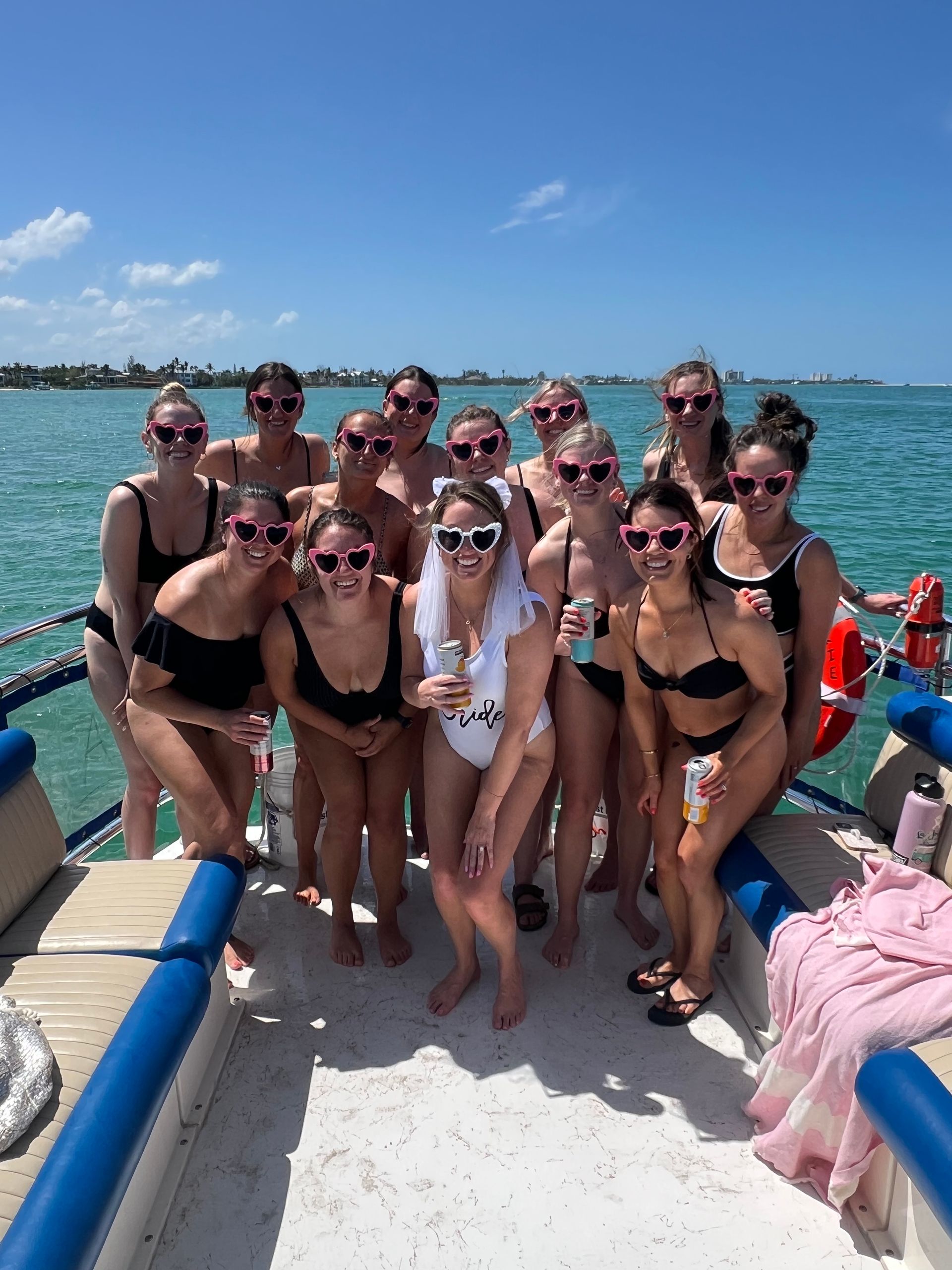 Bachelorette party on a boat: group of women in swimsuits wearing pink heart-shaped sunglasses, bride in a white swimsuit and veil, holding drinks on turquoise coastal water under a sunny blue sky.