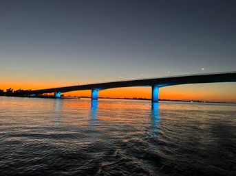 Sunset waterfront bridge with glowing blue-lit pillars reflecting across calm water under an orange-to-deep-blue sky