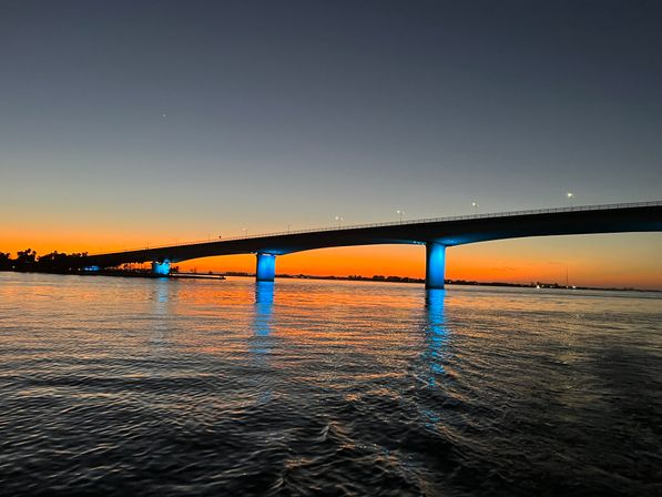 Sunset waterfront bridge with glowing blue-lit pillars reflecting across calm water under an orange-to-deep-blue sky
