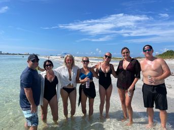Group of seven adults in swimsuits standing waist‑deep in clear shallow water on a sunny sandy beach, smiling under a bright blue sky with umbrellas and distant shoreline — casual summer beach day with drinks.