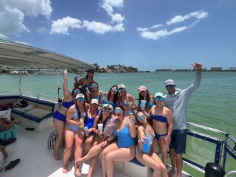 Sunlit boat party with a cheerful group in swimsuits and mirrored sunglasses holding drinks on a pontoon in turquoise coastal waters, palm-lined shore and blue sky in the background.