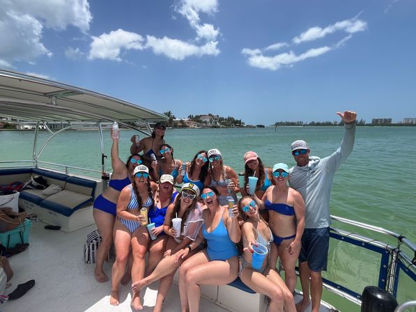 Sunlit boat party with a cheerful group in swimsuits and mirrored sunglasses holding drinks on a pontoon in turquoise coastal waters, palm-lined shore and blue sky in the background.