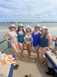 Five friends in swimsuits and straw hats posing on a pontoon boat by a sandy shoreline with a coastal skyline and cloudy sky in the background — sunny beach day on the bay.