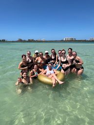 Group of friends at a sunny beach bachelorette party in shallow turquoise water, women in swimsuits and heart-shaped sunglasses surrounding the bride lounging on a gold inflatable ring with resort buildings on the distant shoreline.