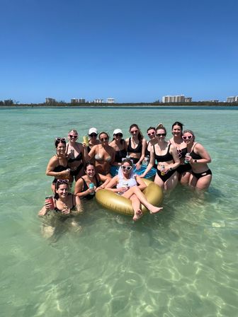Group of friends at a sunny beach bachelorette party in shallow turquoise water, women in swimsuits and heart-shaped sunglasses surrounding the bride lounging on a gold inflatable ring with resort buildings on the distant shoreline.