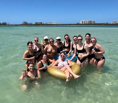 Group of friends at a sunny beach bachelorette party in shallow turquoise water, women in swimsuits and heart-shaped sunglasses surrounding the bride lounging on a gold inflatable ring with resort buildings on the distant shoreline.