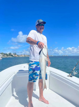 Smiling angler on a white boat holding a large silver fish over coastal blue water and clear sky, wearing sunglasses, cap and blue camo shorts — recreational saltwater fishing scene.