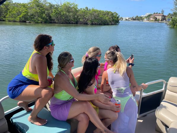 Group of friends in colorful swimwear on a pontoon boat taking a selfie and holding canned drinks on a sunny mangrove-lined coastal inlet.